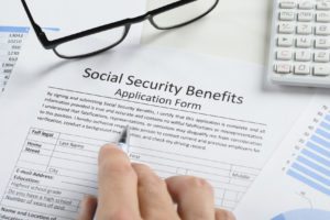 Close-up Of Hand With Pen And Eyeglasses Over Social Security Benefits Application Form